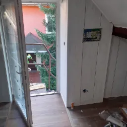 Poddasze w trakcie wykańczania: białe panele ścienne imitujące drewno, otwarte drzwi balkonowe z widokiem na zieleń, widoczne kable elektryczne i narzędzia pracy.