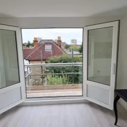 Wnętrze pokoju z otwartymi dwuskrzydłowymi białymi drzwiami balkonowymi, widok na balkon z metalową barierką i zabudowę miejską w tle.