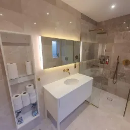 Elegancka, wykończona łazienka w Łętowicach z marmurowymi płytkami, podświetlanym lustrem nad białą umywalką i kabiną prysznicową typu walk-in z złotymi akcentami.