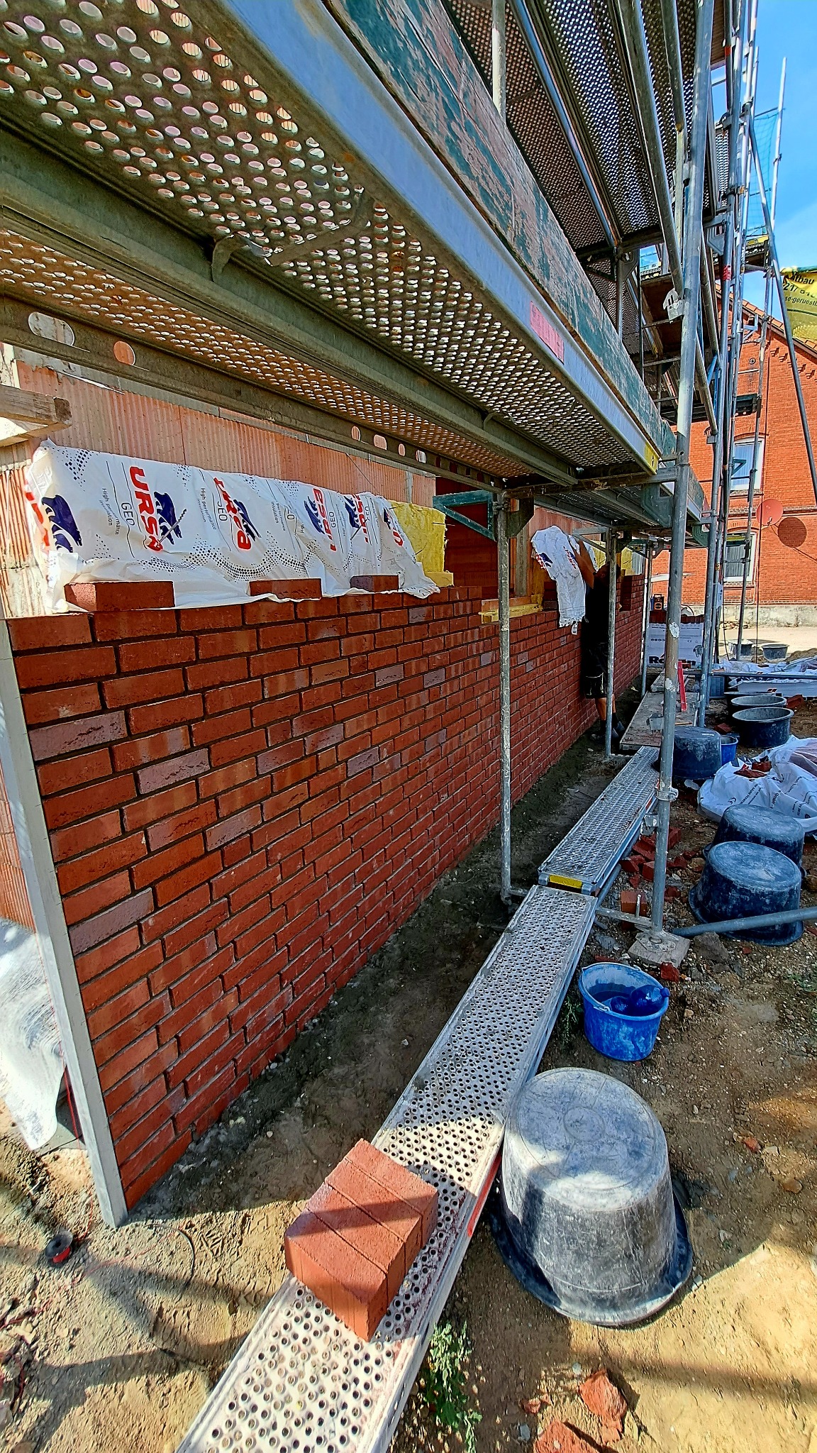 Świeżo wymurowana ściana z czerwonej cegły pod rusztowaniem, z widocznym ociepleniem i materiałami budowlanymi, w trakcie prac wykończeniowych na zewnątrz budynku.