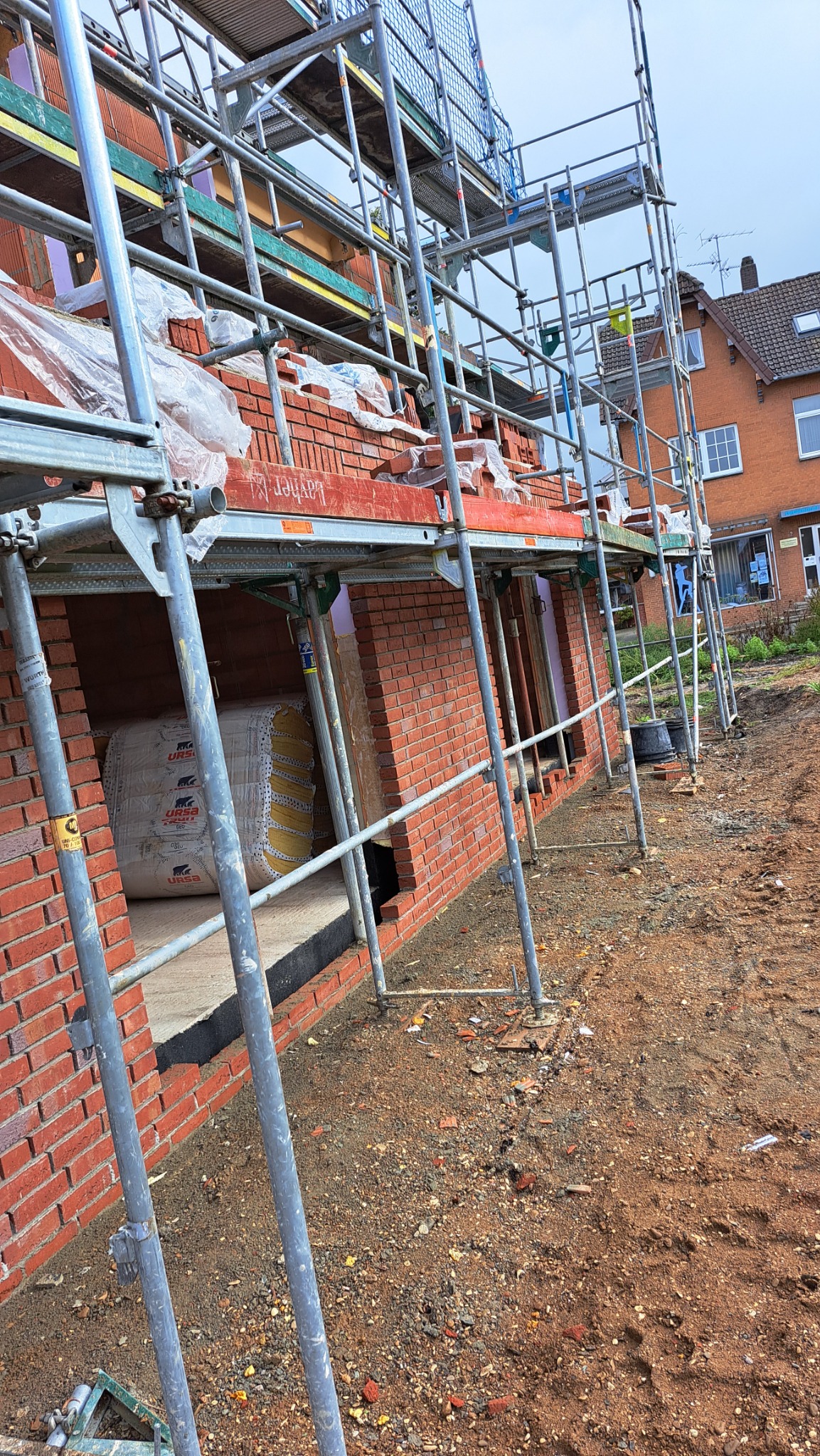Budowa domu w Weeze: ściany z czerwonej cegły w trakcie murowania, widoczne rusztowania i rolka materiału izolacyjnego URSA.