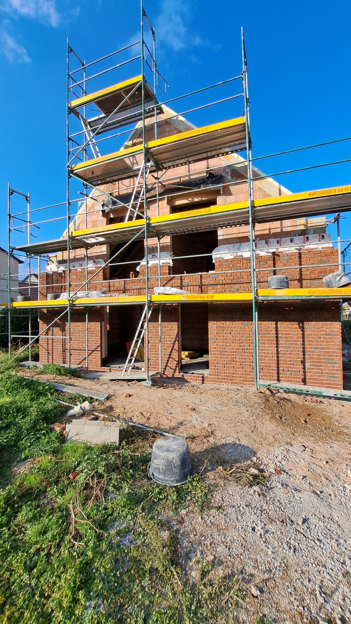Budowa domu jednorodzinnego z czerwonej cegły, z widocznymi rusztowaniami i częściowo wzniesionymi ścianami, na tle błękitnego nieba.