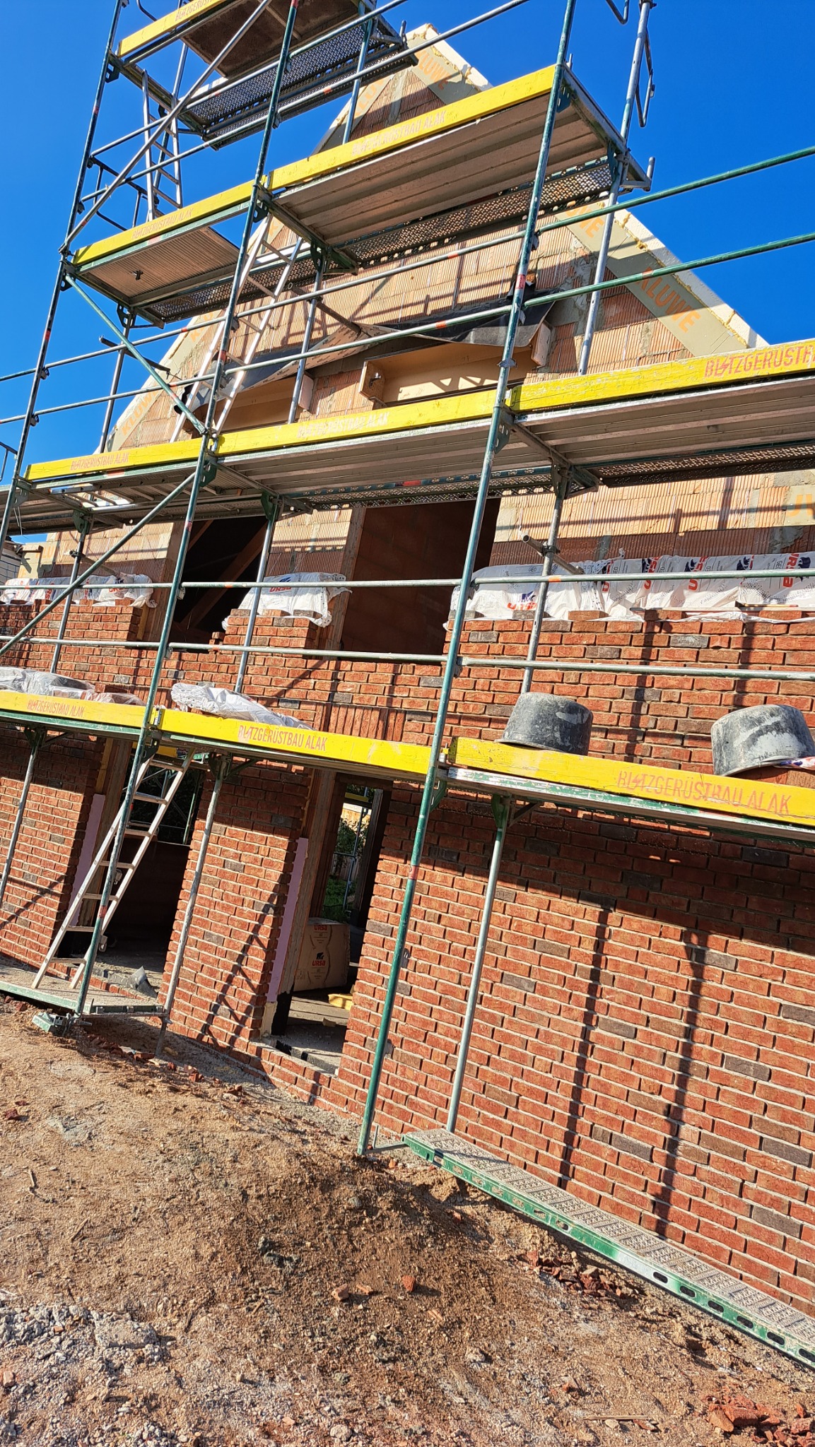 Wznoszenie muru z czerwonej cegły na nowym budynku, widoczne rusztowanie, zaprawa, pustaki ceramiczne i ocieplenie w szczycie dachu.