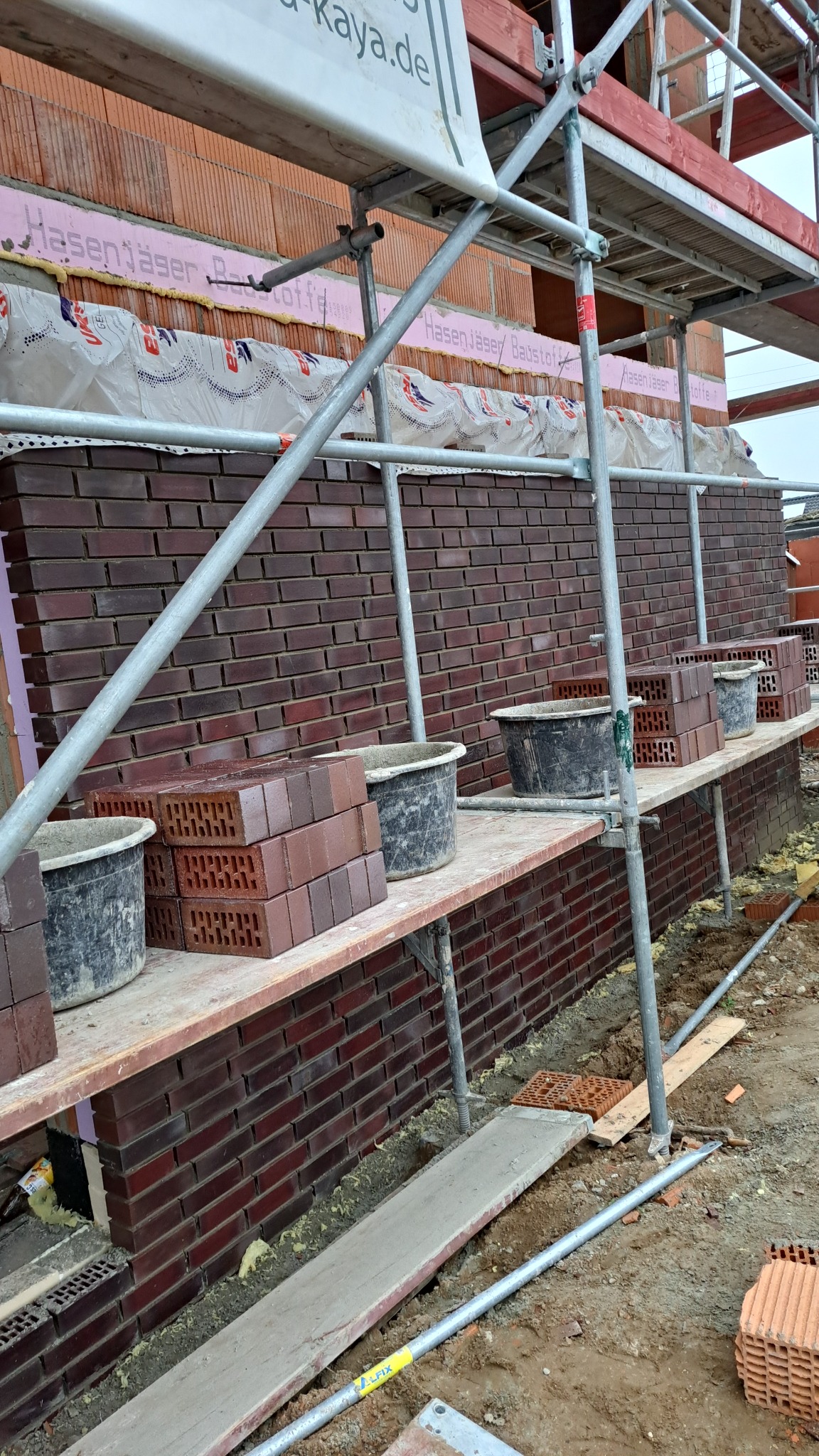 Murowana ściana z czerwonej cegły na budowie, widoczne rusztowanie, zapas cegieł i wiadra z zaprawą na drewnianych deskach, ocieplenie ściany widoczne w górnej części.