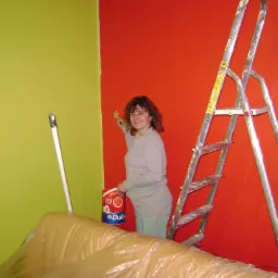 Kobieta uśmiecha się, malując czerwoną ścianę pędzlem, obok drabina i wiadro farby Dulux, część ściany pomalowana na zielono, podłoga zabezpieczona folią malarską.