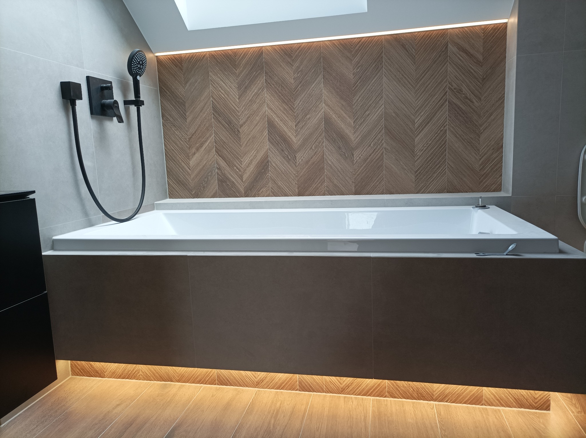 Nowoczesna łazienka z białą wanną, armaturą w kolorze czarnym i podświetleniem LED pod wanną i nad dekoracyjną ścianą z drewnopodobnych płytek ułożonych w jodełkę.