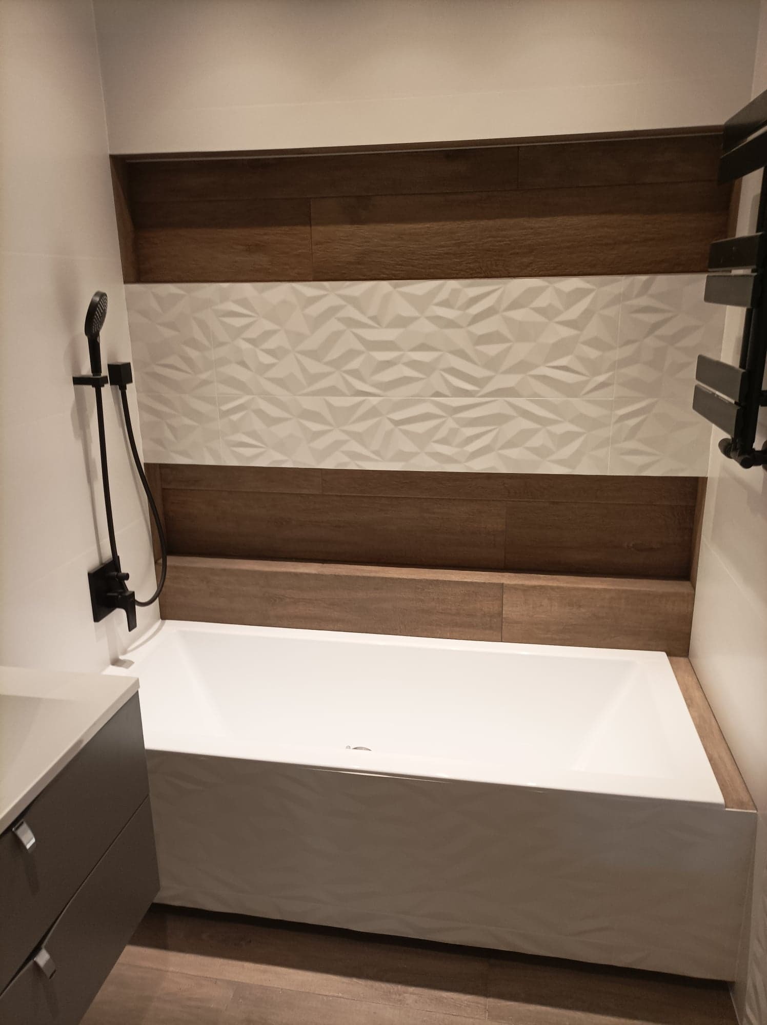 Nowoczesna łazienka z białą wanną obudowaną płytkami imitującymi drewno, ściana wykończona białymi płytkami 3D i pasami dekoracyjnymi z płytek drewnopodobnych, czarna armatura.