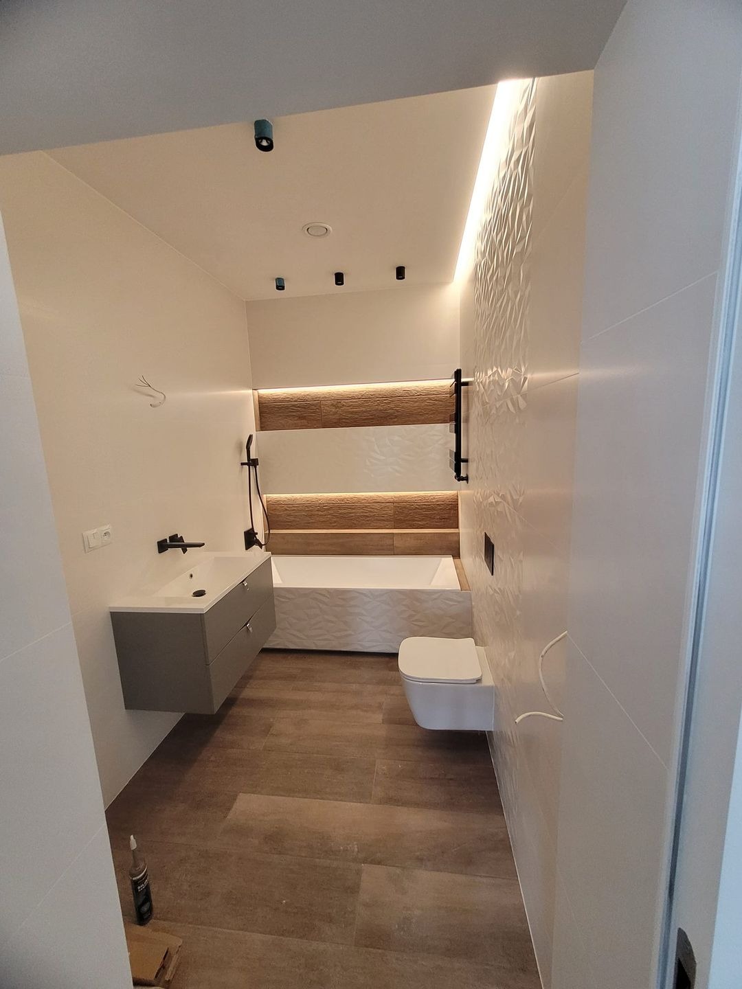 Nowoczesna łazienka z drewnopodobną podłogą, białą armaturą, geometryczną ścianą, szarą szafką pod umywalką i oświetleniem LED.