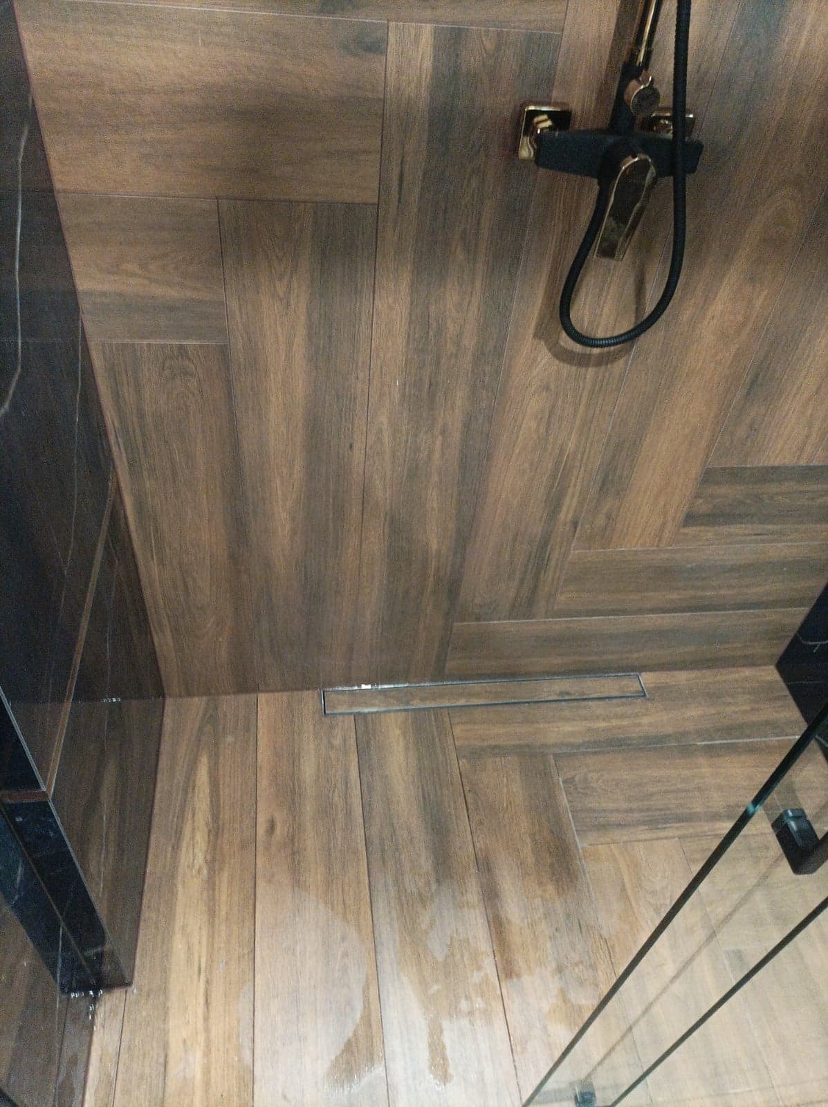 Ukośny widok na podłogę prysznica wyłożoną płytkami imitującymi drewno, z odpływem liniowym i czarną armaturą w złotym odcieniu.