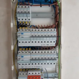 Otwarta skrzynka elektryczna z zainstalowanymi bezpiecznikami Hager i przewodami w różnych kolorach, widoczne także zabezpieczenia przeciwprzepięciowe Simtec z oznaczeniami L1, L2, L3, N i PE.