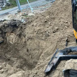 Wykop w ziemi zabezpieczony ogrodzeniem tymczasowym, widoczny fragment koparki i sterta ziemi. W tle budynek z czerwonej cegły.