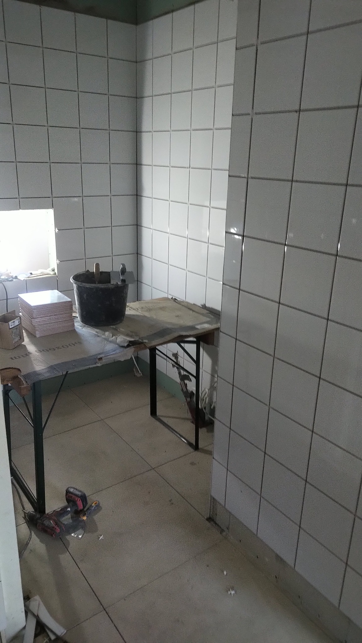 Proces układania białych płytek ceramicznych na ścianie w łazience, widoczny stół roboczy z narzędziami i wiaderkiem z zaprawą.