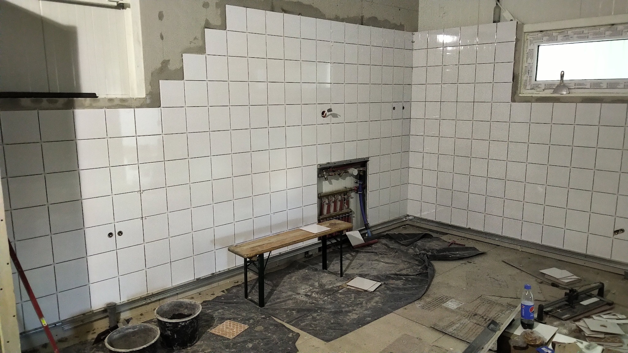 Wnętrze w trakcie układania białych, kwadratowych płytek na ścianach, widoczne instalacje hydrauliczne, stół roboczy i narzędzia.