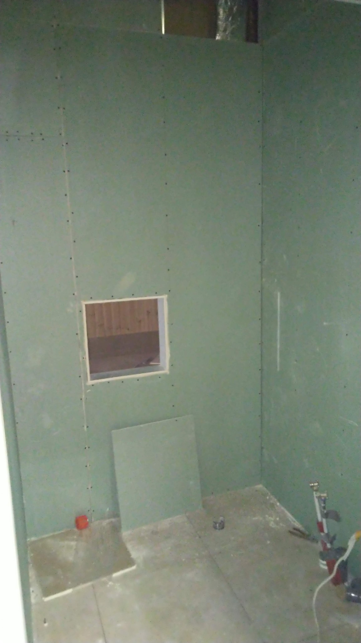 Wnętrze w trakcie wykańczania, ściany pokryte zielonymi płytami gipsowo-kartonowymi, otwór okienny z widoczną drewnianą konstrukcją w środku, narzędzia budowlane leżące na podłodze.