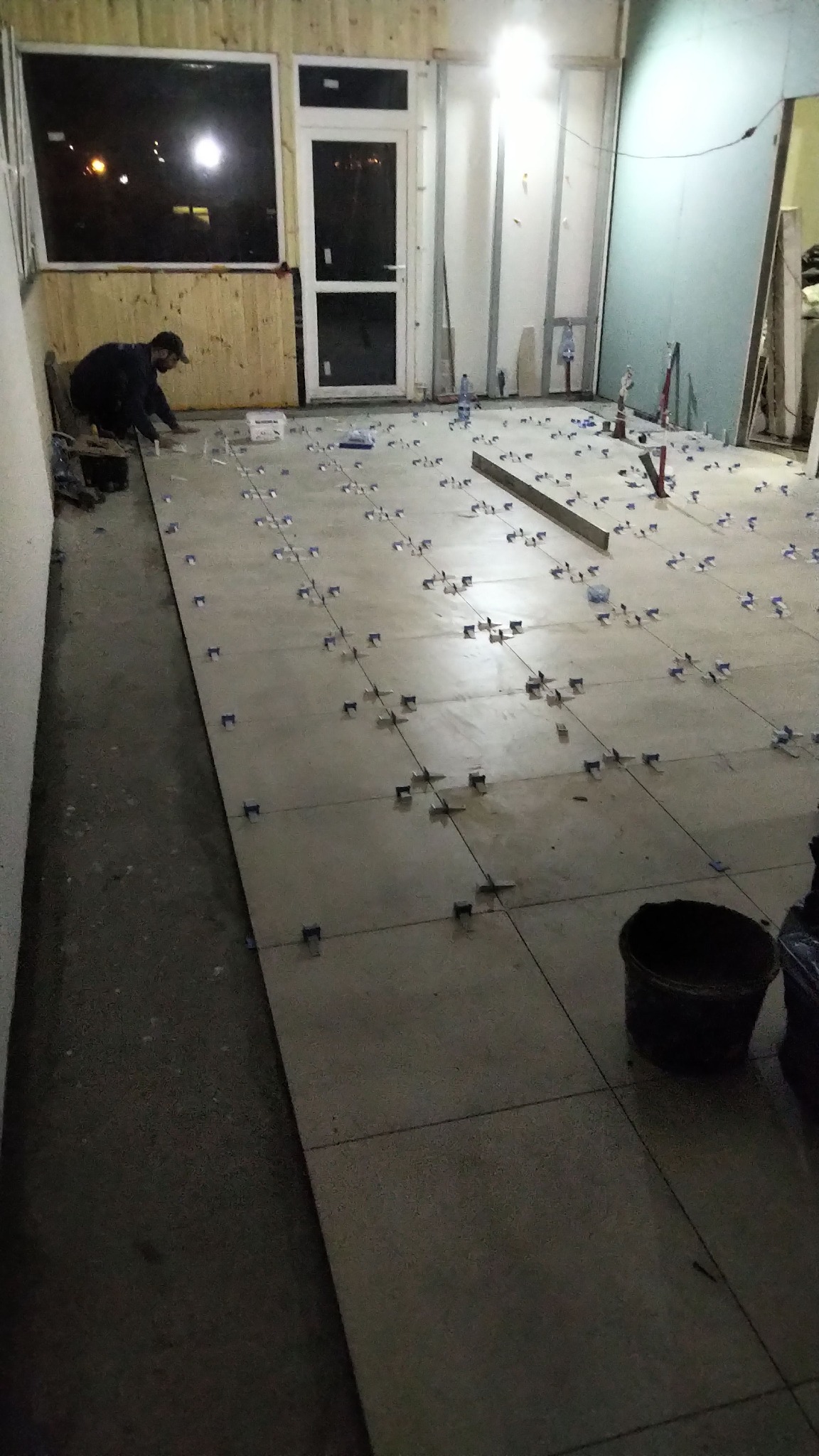 Układanie szarych płytek podłogowych z użyciem krzyżyków dystansowych, widoczny pracownik w masce, poziomica i wiadro