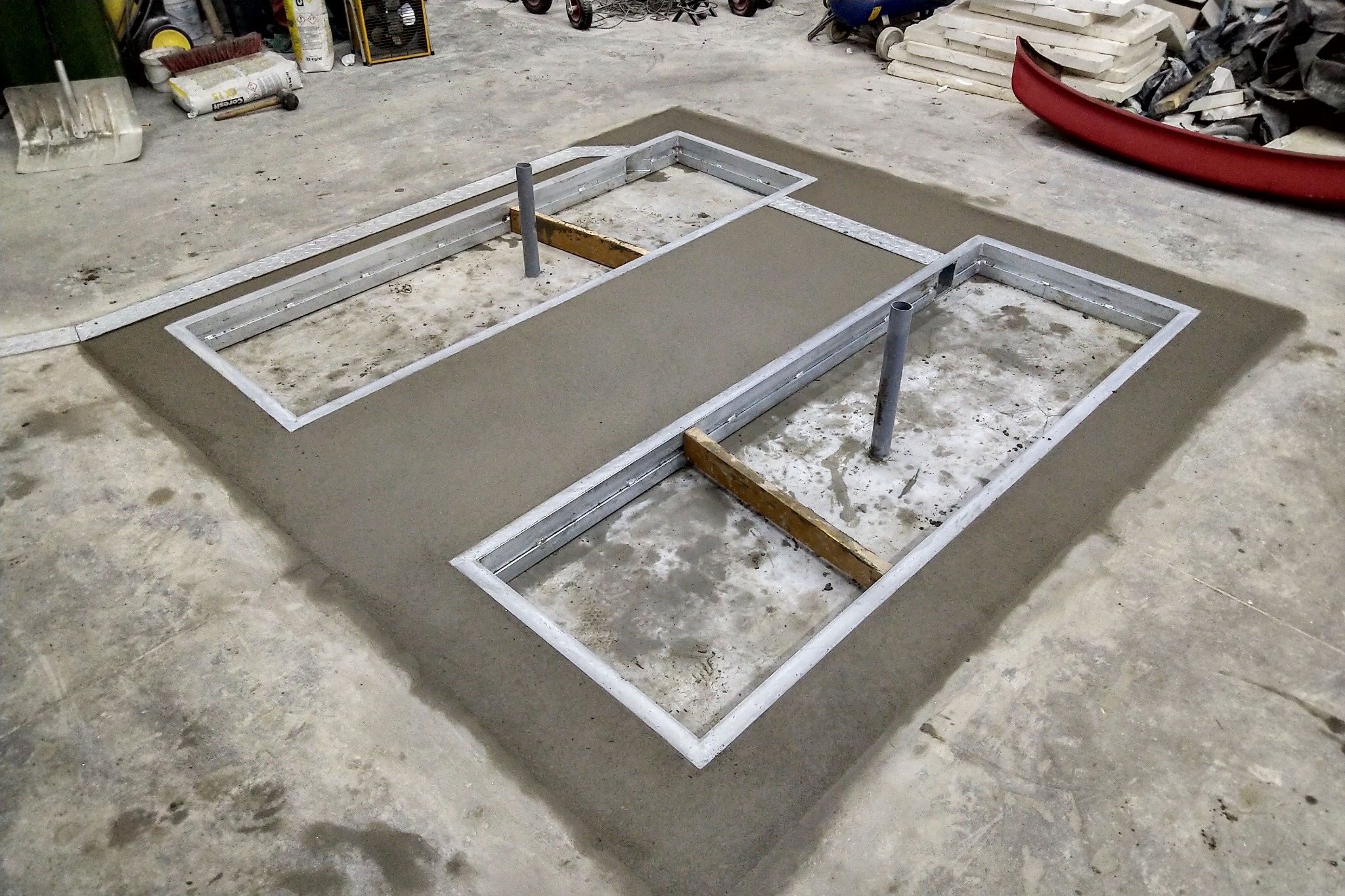 Przygotowanie podłoża pod wylewkę betonową z metalowymi ramami i wystającymi rurami, widoczne narzędzia i materiały budowlane w tle.