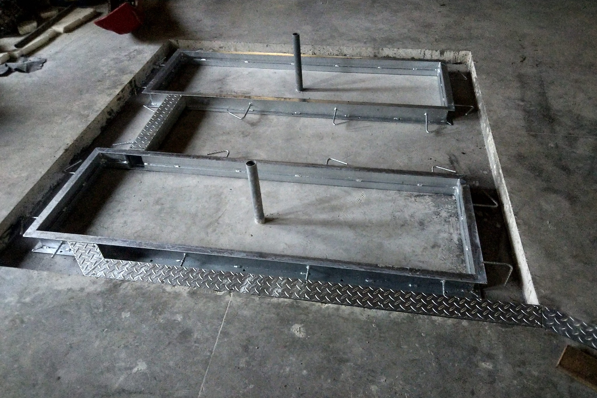 Dwie metalowe ramy z wystającymi rurami, osadzone w betonowej posadzce z elementami antypoślizgowymi, przygotowane do dalszych prac konstrukcyjnych.