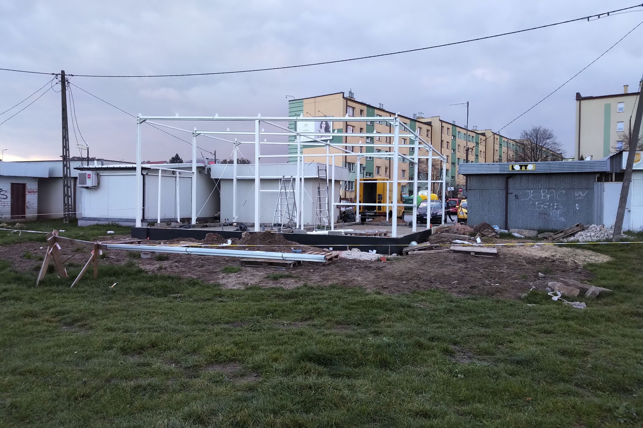 Konstrukcja stalowa szkieletu budynku w trakcie montażu na przygotowanym fundamencie, widoczne rusztowanie i elementy zabezpieczające teren budowy.