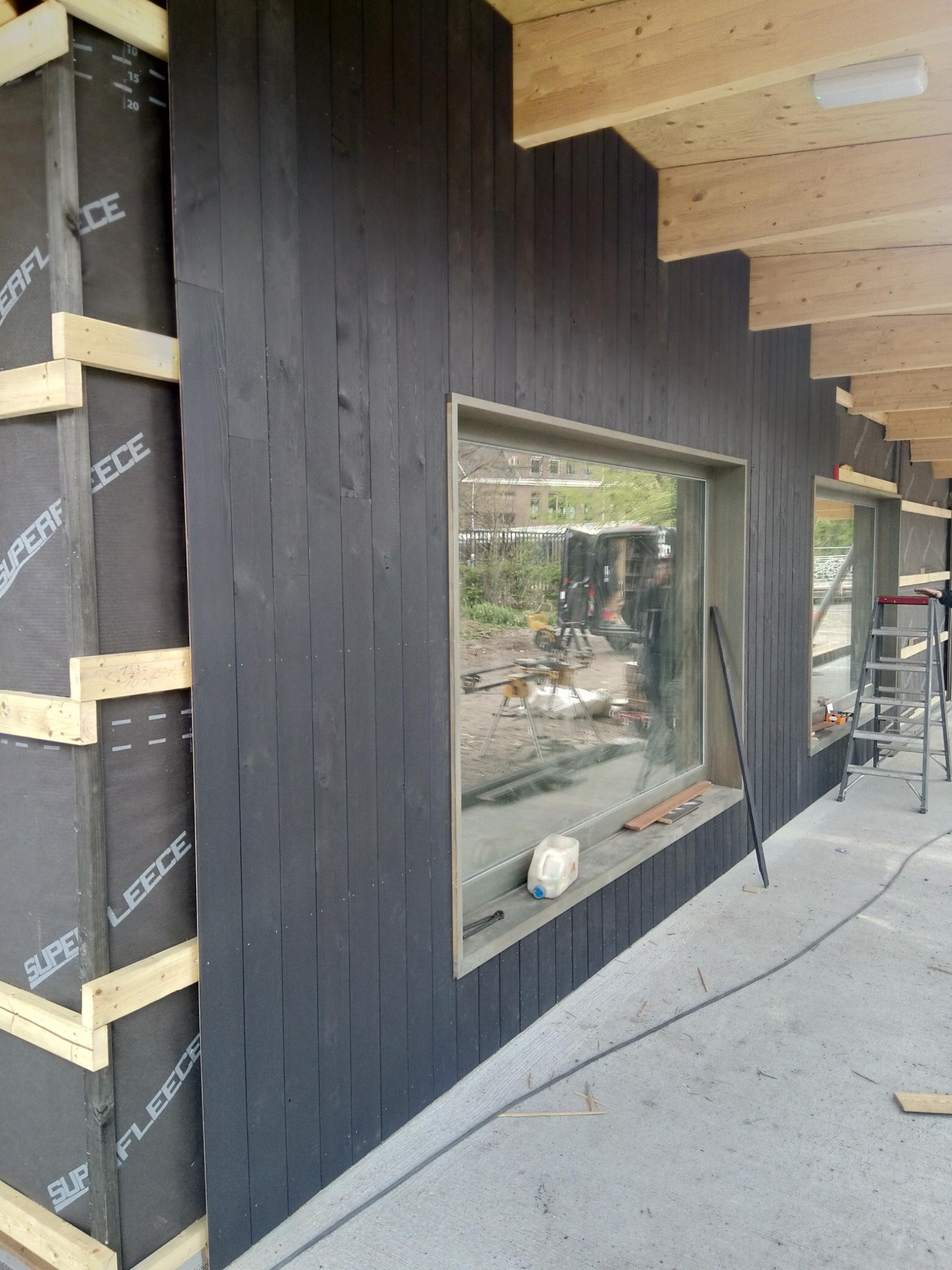 Montaż elewacji z ciemnego drewna, widoczna izolacja i fragment więźby dachowej, okno z odbiciem otoczenia