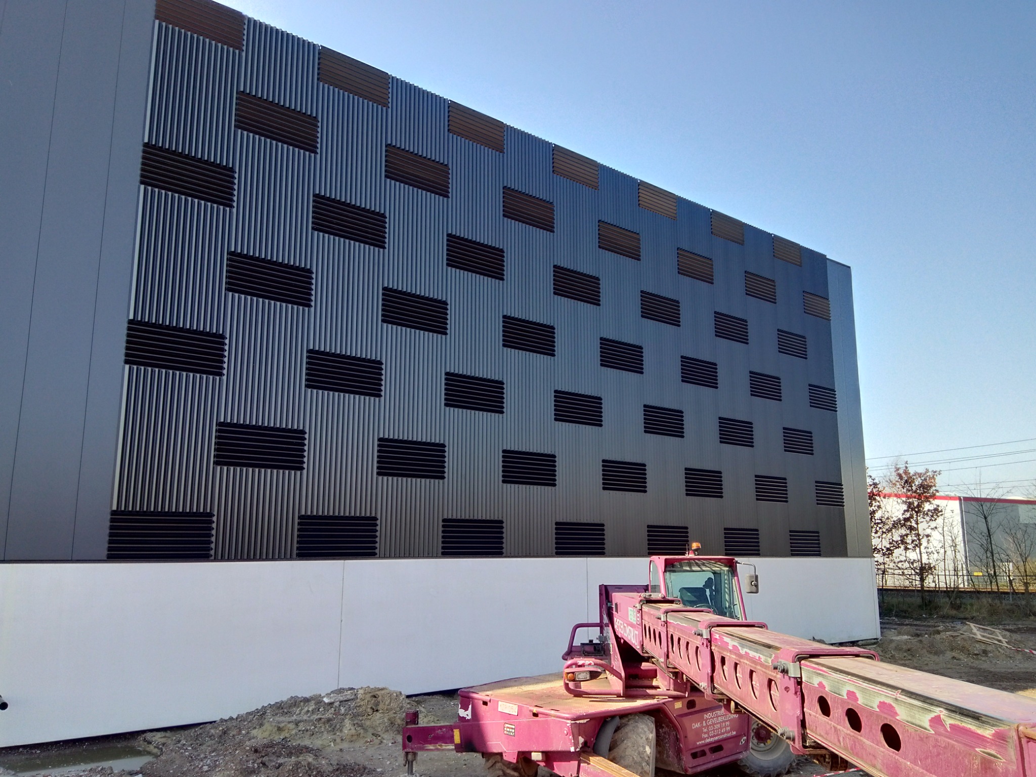 Montaż elewacji wentylowanej z metalowych paneli z poziomymi przesłonami wentylacyjnymi, widoczny podnośnik teleskopowy na placu budowy.