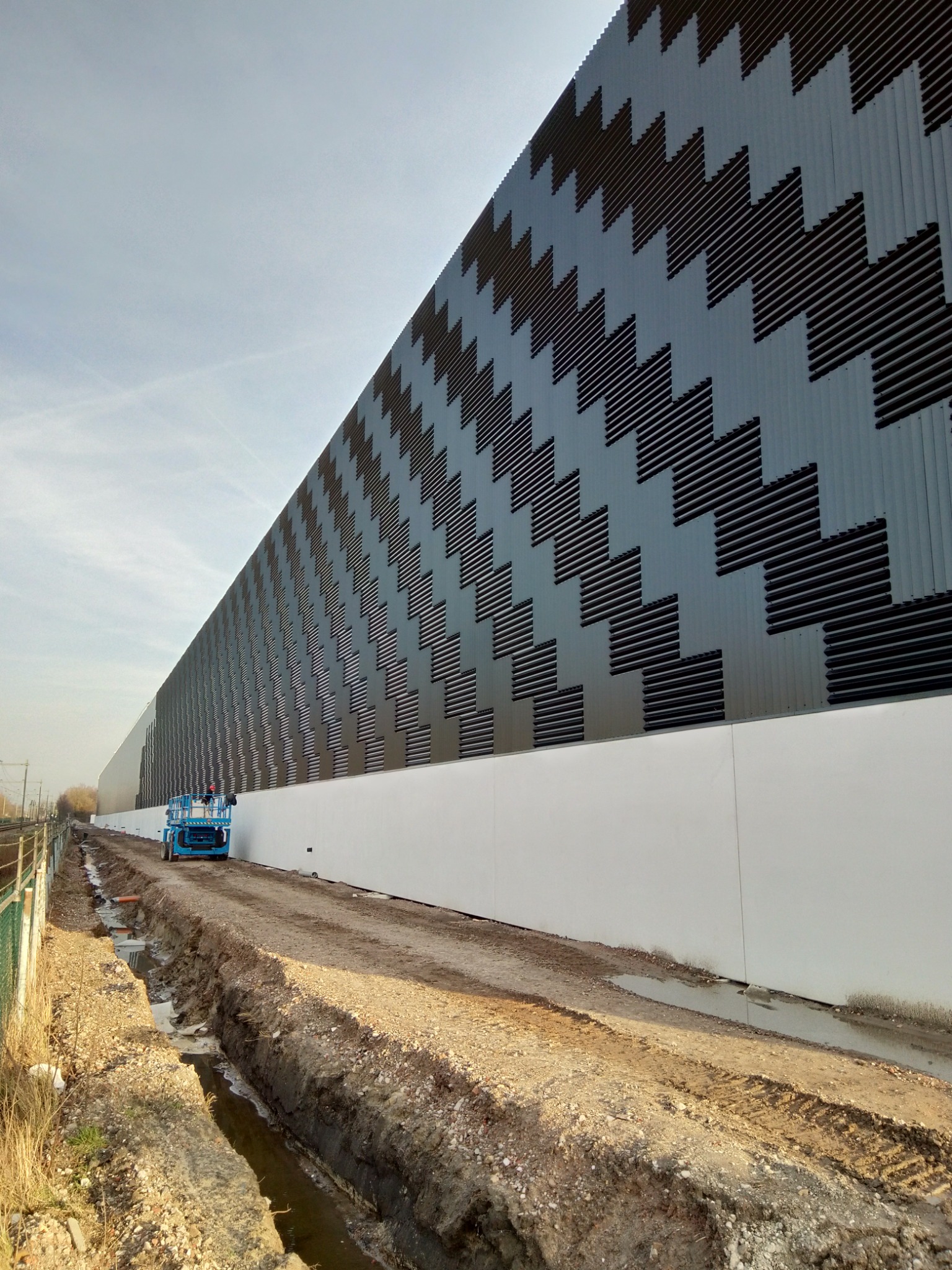 Montaż elewacji wentylowanej z wzorem zygzakowatym na wysokim budynku przemysłowym, widoczna podnośnik koszowy i teren budowy.