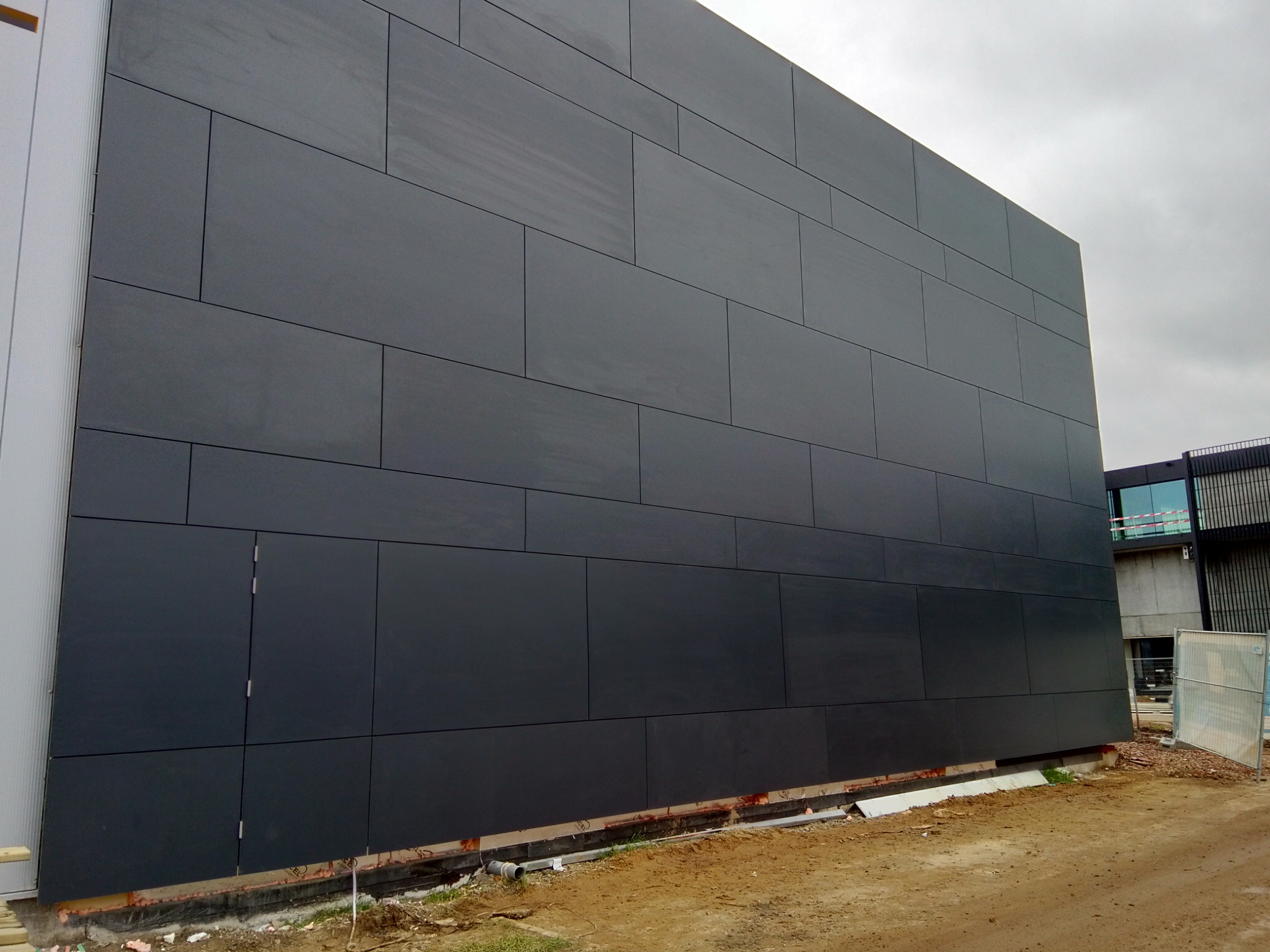 Elewacja budynku wykończona panelami kompozytowymi w kolorze grafitowym, widoczne fragmenty mocowań i otwór rewizyjny w dolnej części ściany, teren wokół w trakcie prac ziemnych.