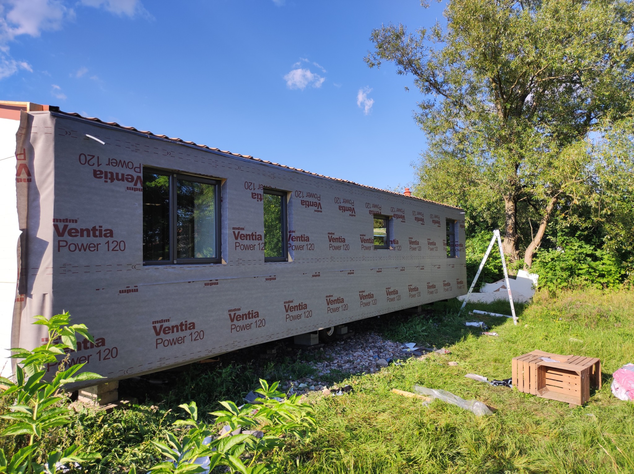 Modułowy dom w trakcie budowy, zabezpieczony folią wiatroizolacyjną Ventia Power 120, widoczne okna i fragment dachu, otoczenie zielone z drzewami.