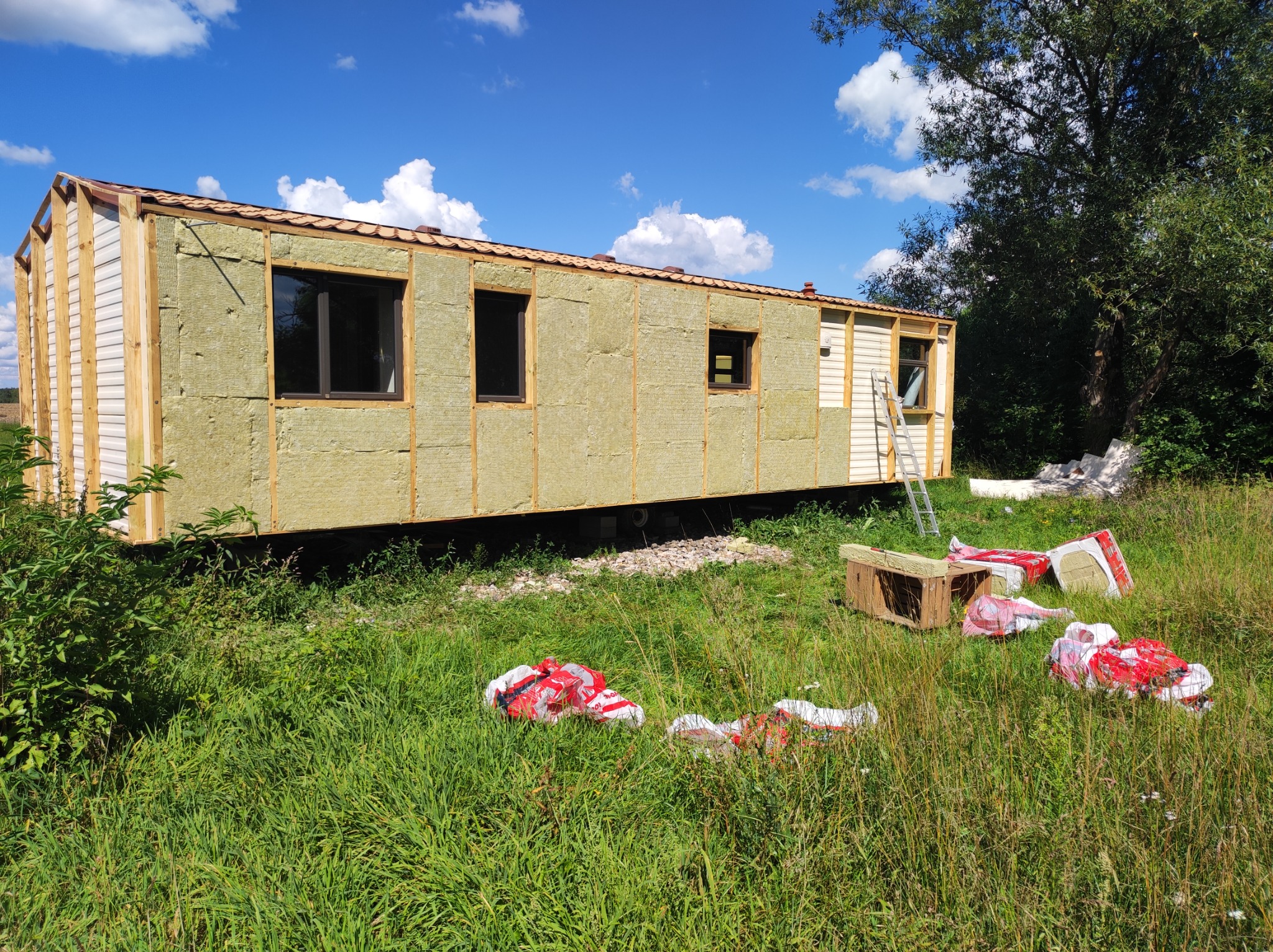 Budowa szkieletowego domu mobilnego z izolacją, częściowo obłożonego sidingiem, na działce z wysoką trawą i rozpakowanymi paczkami wełny mineralnej.