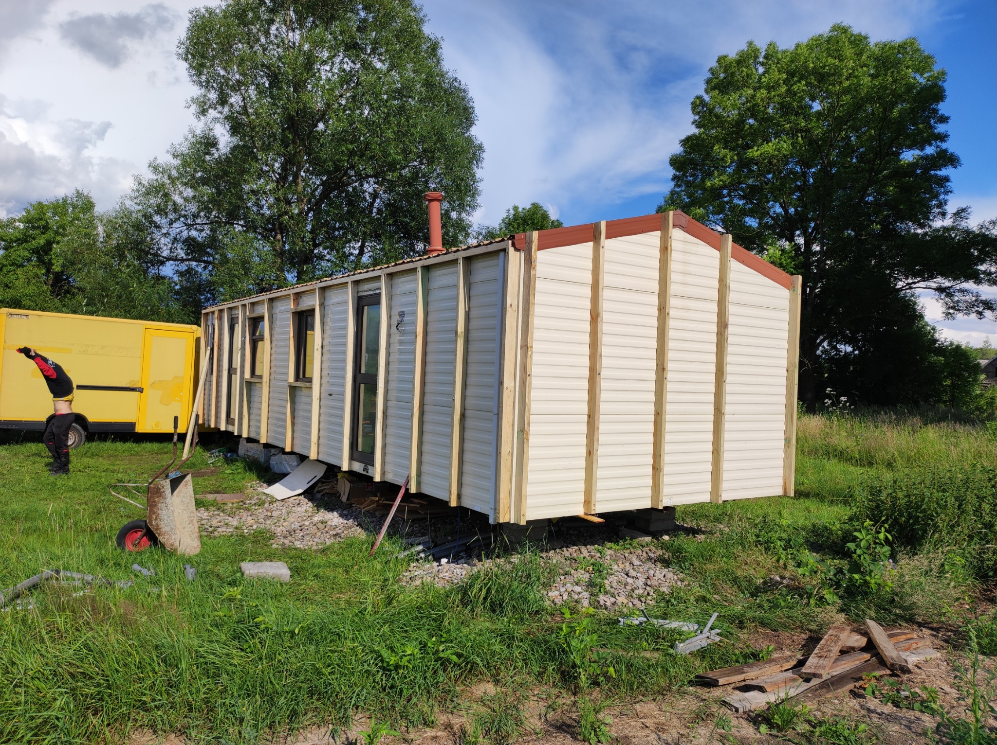 Montaż obudowy domku letniskowego z panelami sidingowymi i drewnianą konstrukcją szkieletową, widoczny wózek budowlany i teren zielony.