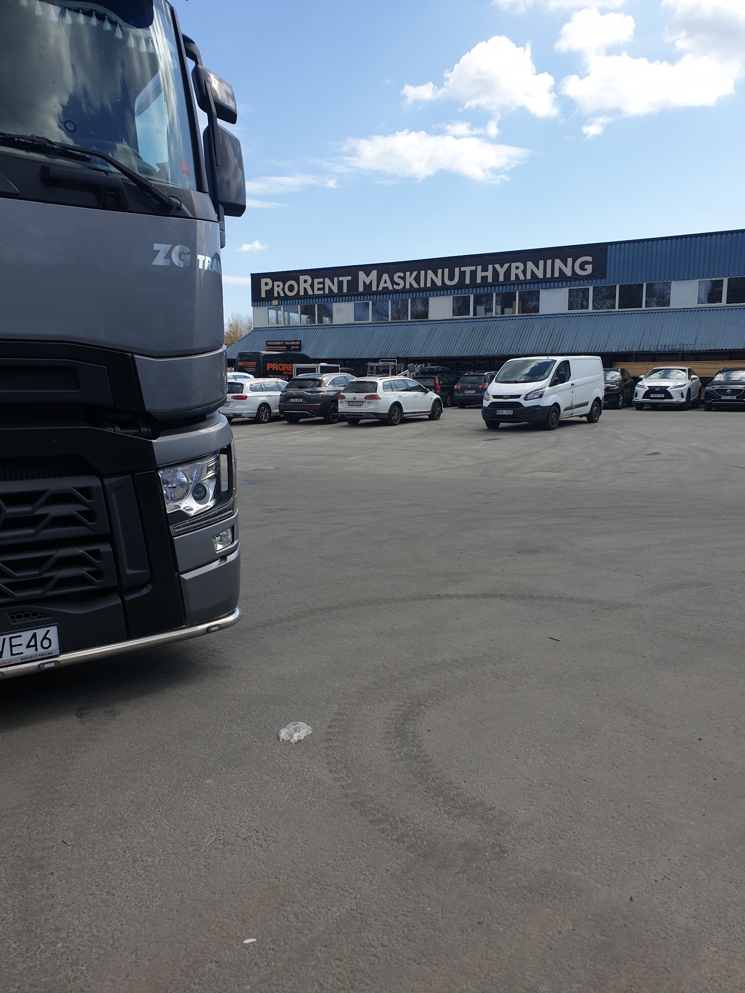 Częściowy widok szarej ciężarówki ZG Trans na parkingu przed budynkiem Prorent Maskinuthyrning, z zaparkowanymi samochodami osobowymi i dostawczymi w tle.