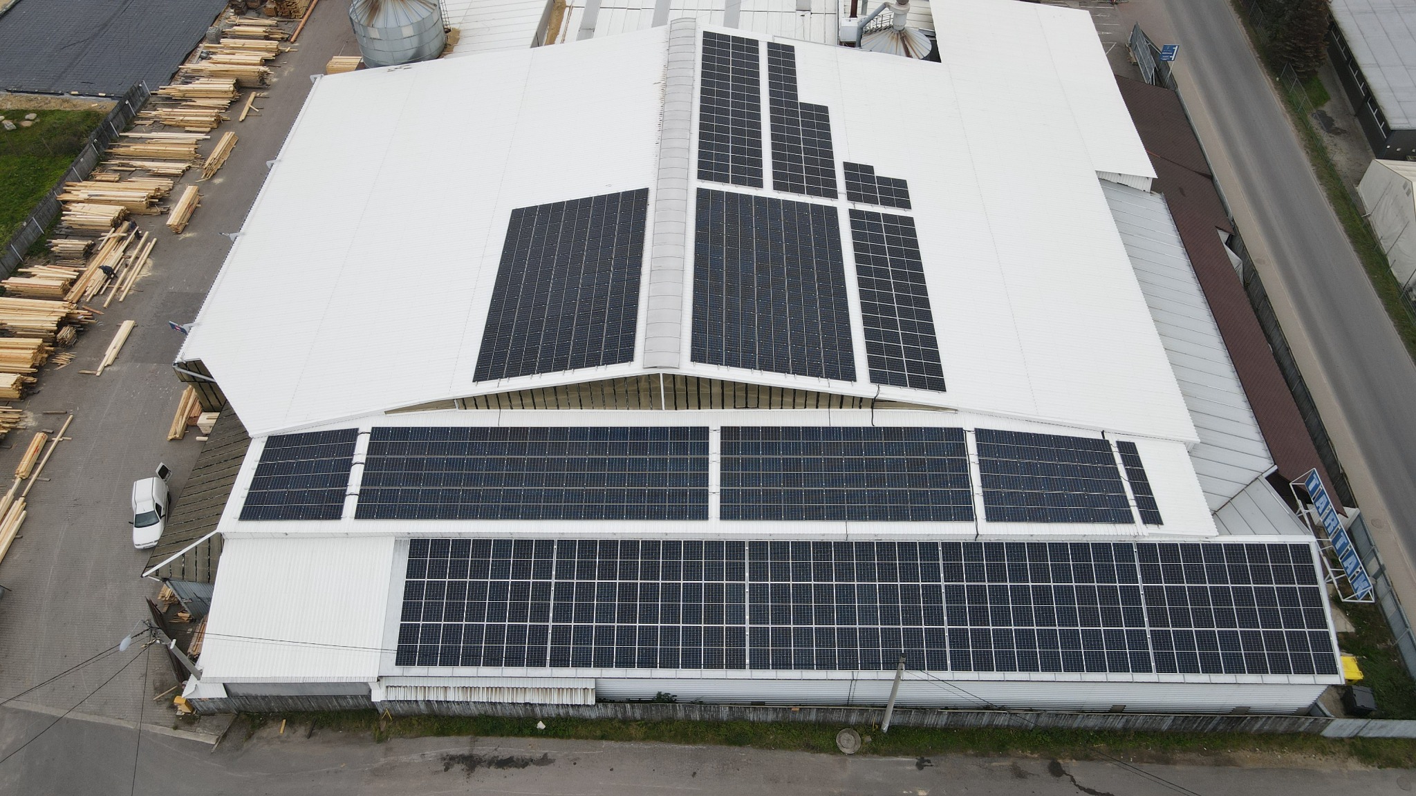 Widok z góry na biały dach hali przemysłowej z zamontowanymi panelami słonecznymi w układzie blokowym, obok składowane drewno i zaparkowany samochód dostawczy.