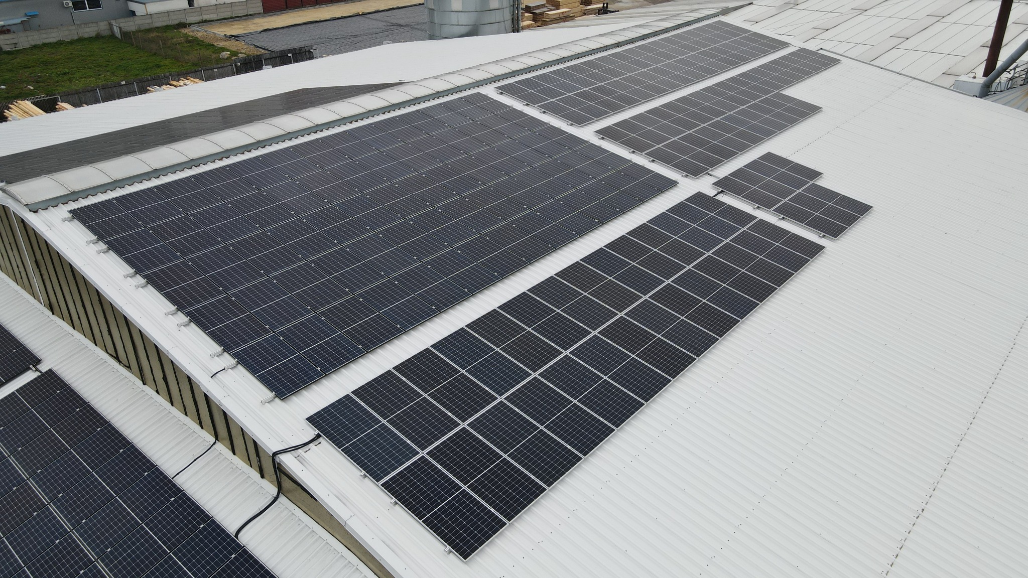 Widok z góry na instalację paneli słonecznych na dachu przemysłowego budynku z blachy falistej. Panele ułożone w rzędach, różnej wielkości, z widocznymi przewodami.