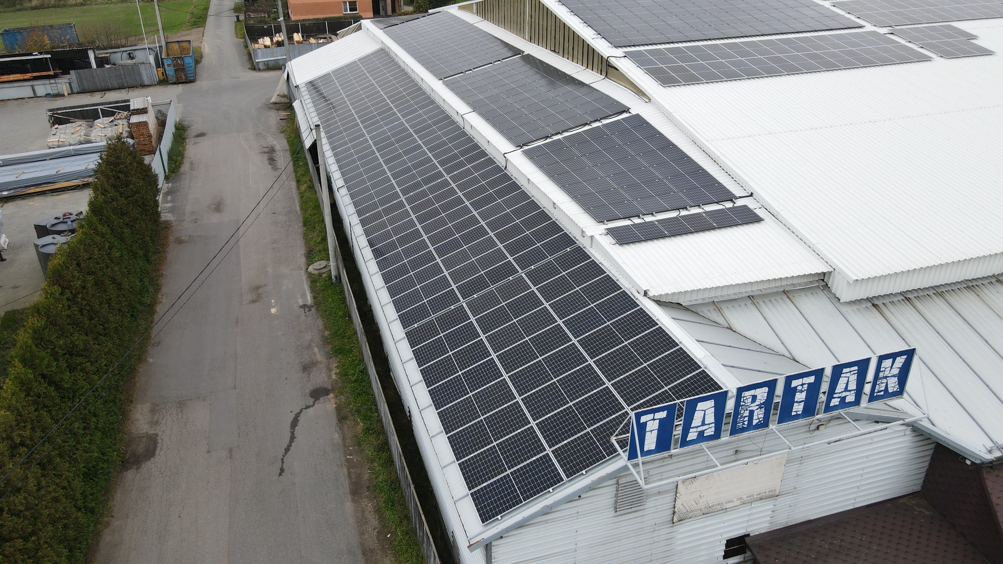 Widok z góry na dużą instalację fotowoltaiczną na dachu budynku przemysłowego z widocznym napisem 'TARTAK' na elewacji.