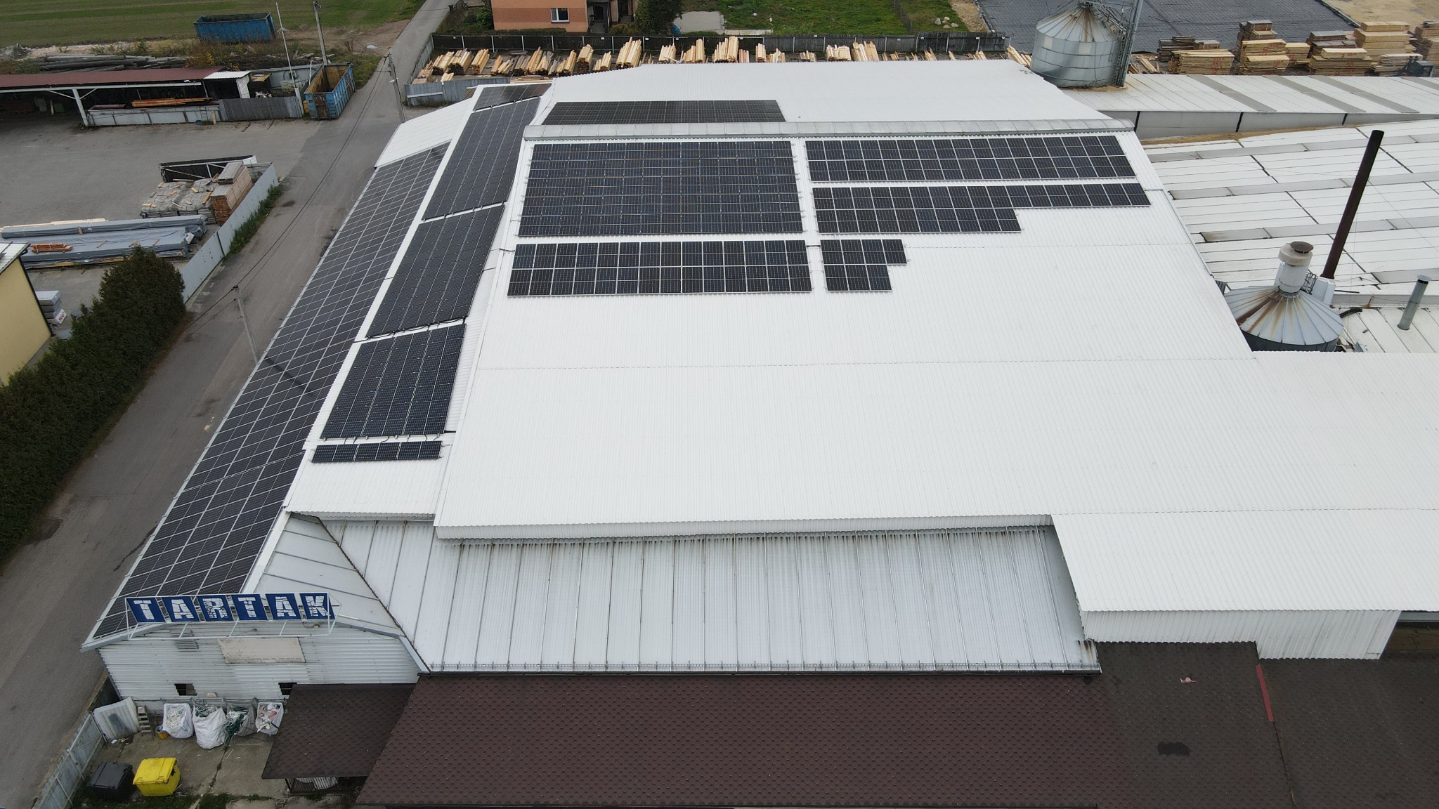 Widok z góry na duży budynek przemysłowy z zainstalowanymi panelami słonecznymi na dachu, obok widoczne silosy i składowane drewno, tablica z nazwą 'TARTAK' na froncie budynku.