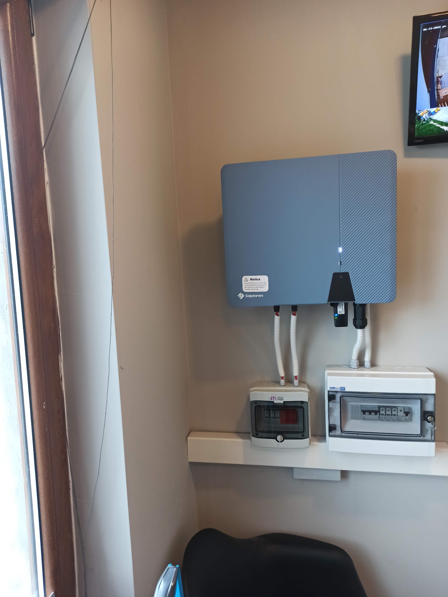 Szary inwerter fotowoltaiczny Solplanet zamontowany na ścianie, z widocznymi przewodami i dwoma skrzynkami z bezpiecznikami na półce poniżej. Na ścianie w tle widoczny telewizor.