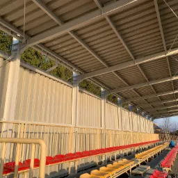 Zadaszenie trybun sportowych z metalową konstrukcją i rzędami czerwono-żółtych siedzeń, widok z perspektywy bocznej w słoneczny dzień.