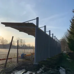 Metalowa konstrukcja z zadaszeniem przy boisku sportowym w zimowej scenerii, widoczne błoto i resztki śniegu, oświetlone porannym słońcem.