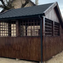 Ciemnobrązowa altana z drewnianą kratą, czarnym dachem z gontu bitumicznego i rynną, na tle drzewa bez liści.