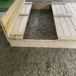 Drewniana konstrukcja z otwieraną klapą, prawdopodobnie piaskownica, na żwirowym podłożu.