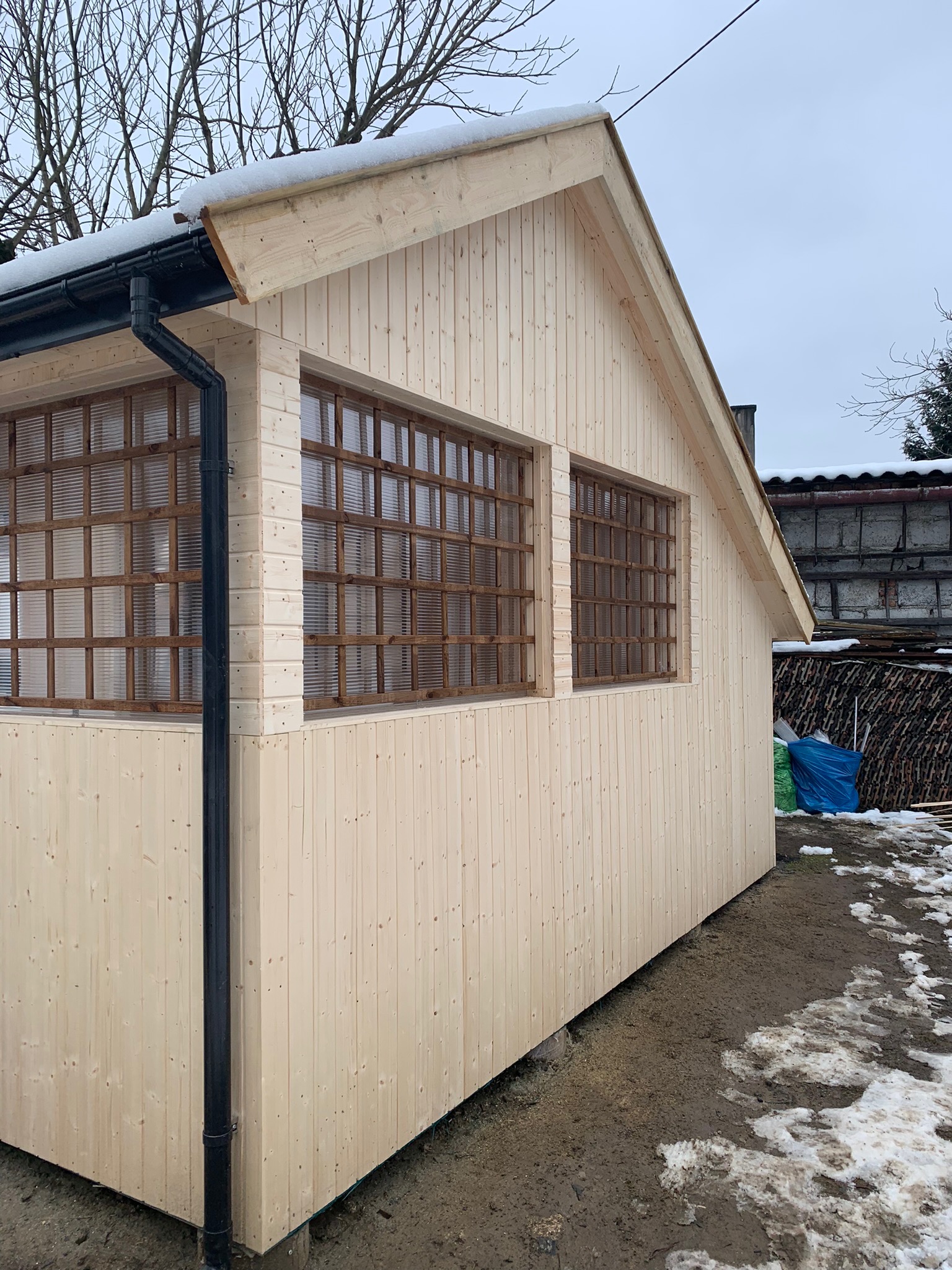 Surowa, drewniana konstrukcja z dwoma oknami z kratownicą, czarną rynną i resztkami śniegu na dachu, stojąca na błotnistym podłożu z topniejącym śniegiem.