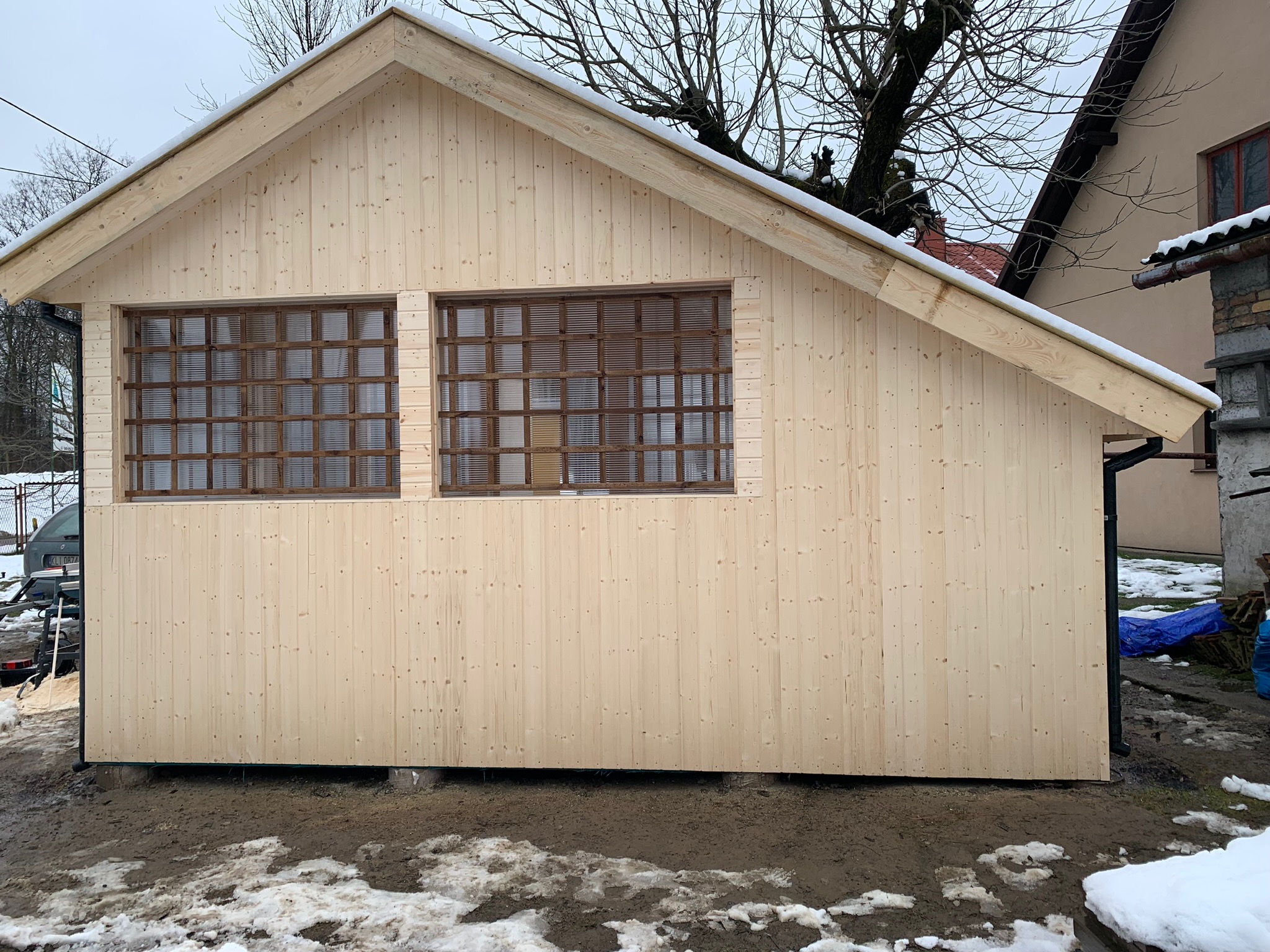 Surowa, drewniana konstrukcja budynku z dwoma oknami z kratownicą, częściowo pokryta śniegiem, na tle błotnistego terenu i zabudowań.