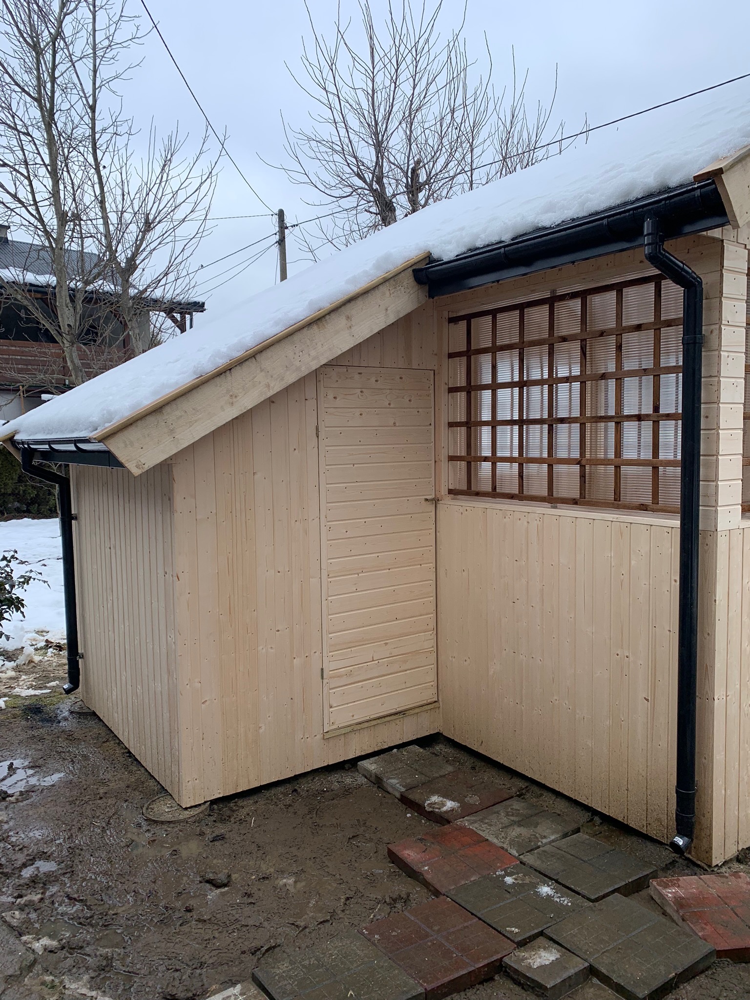 Surowy, drewniany budynek z czarnymi rynnami i kratką okienną, częściowo pokryty śniegiem, na błotnistym podłożu z fragmentami chodnika.