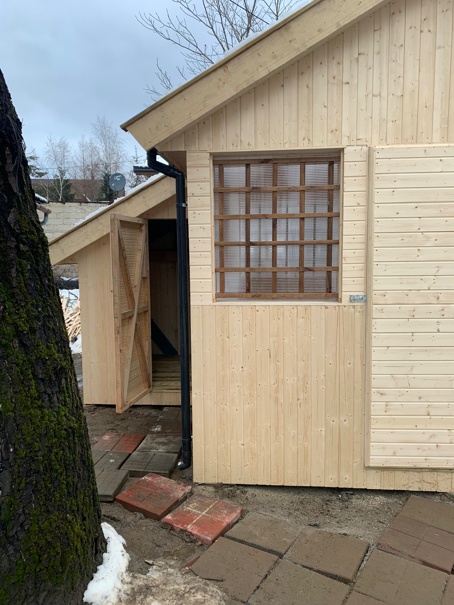 Surowa drewniana konstrukcja z otwartymi drzwiami i oknem z kratką, widoczna na zewnątrz w pochmurny dzień, obok drzewo z mchem i częściowo ułożony chodnik z kostki.