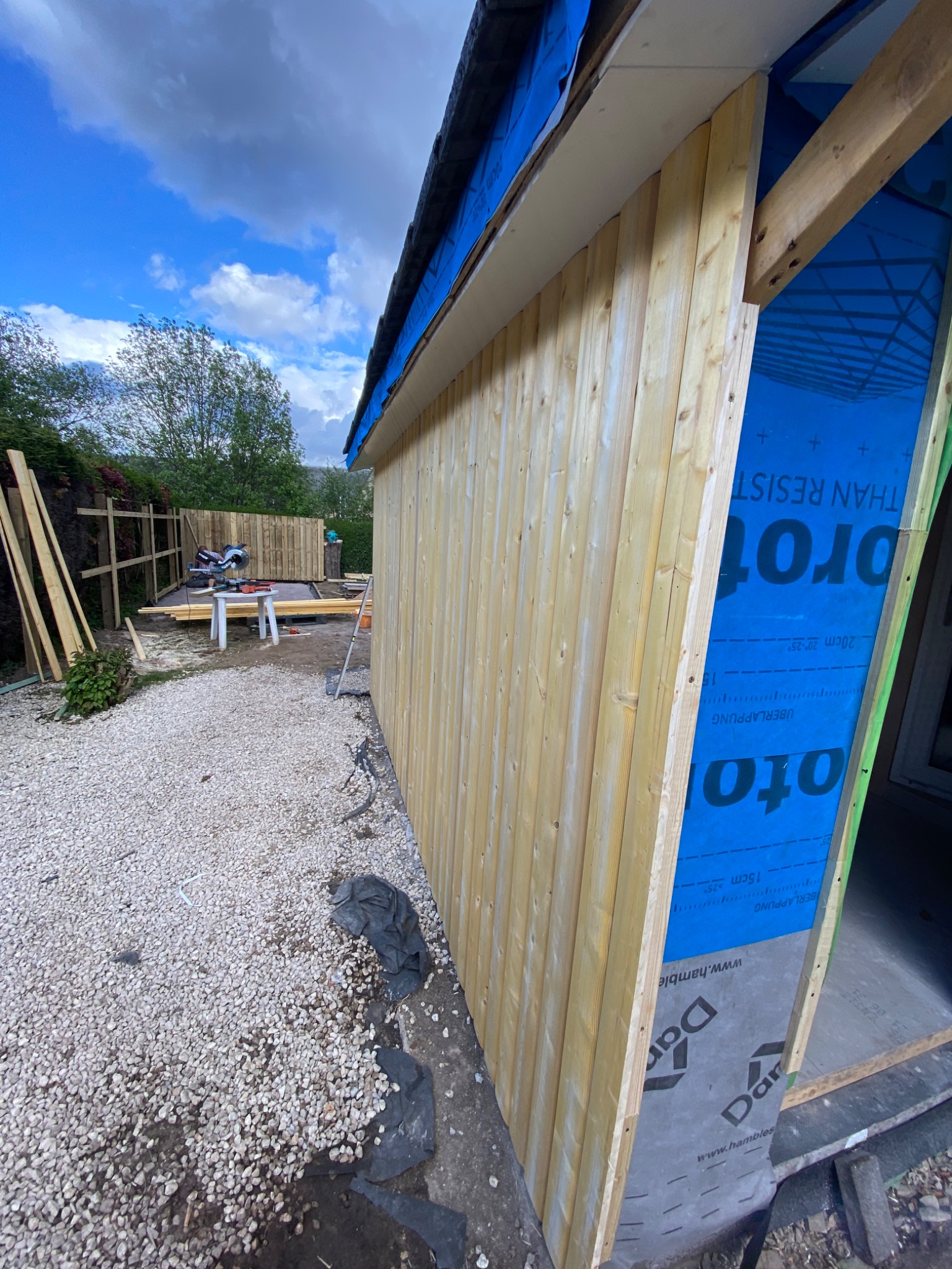 Ściana budynku w trakcie obkładania jasnymi, pionowymi deskami; widoczne narzędzia i materiały budowlane na zewnątrz, niebieska membrana paroizolacyjna.