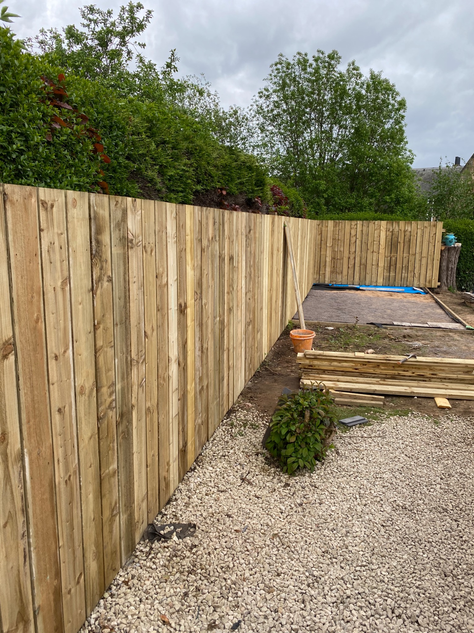 Nowo postawiony, jasny drewniany płot na tle zieleni i szarego nieba, z widocznymi elementami przygotowania terenu i składowanym drewnem.