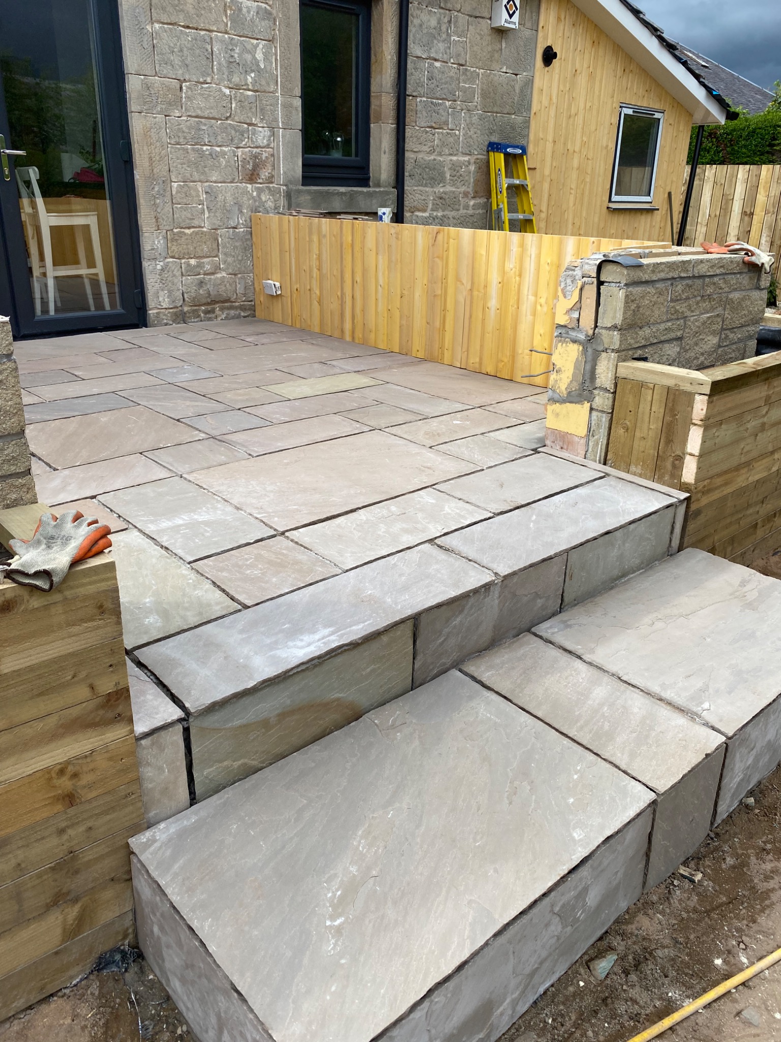Ułożona kamienna kostka brukowa na tarasie i schodach w Glasgow, widoczne drewniane elementy wykończeniowe i rękawice robocze.