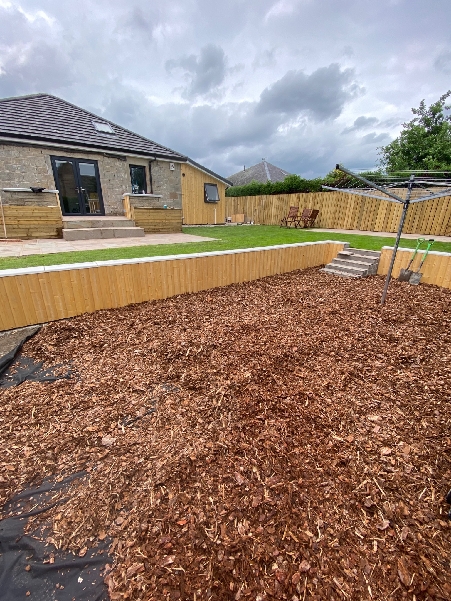 Ukończona aranżacja ogrodu z drewnianym ogrodzeniem, schodami z kostki brukowej i korą rozsypaną na czarnej macie, widoczne narzędzia ogrodnicze i suszarka na pranie.