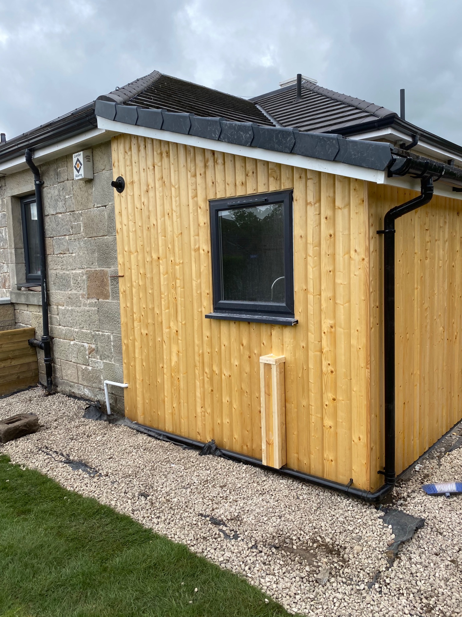 Nowa drewniana dobudówka do kamiennego domu z czarną rynną i oknami, widok z zewnątrz na żwirowe podłoże i trawnik.