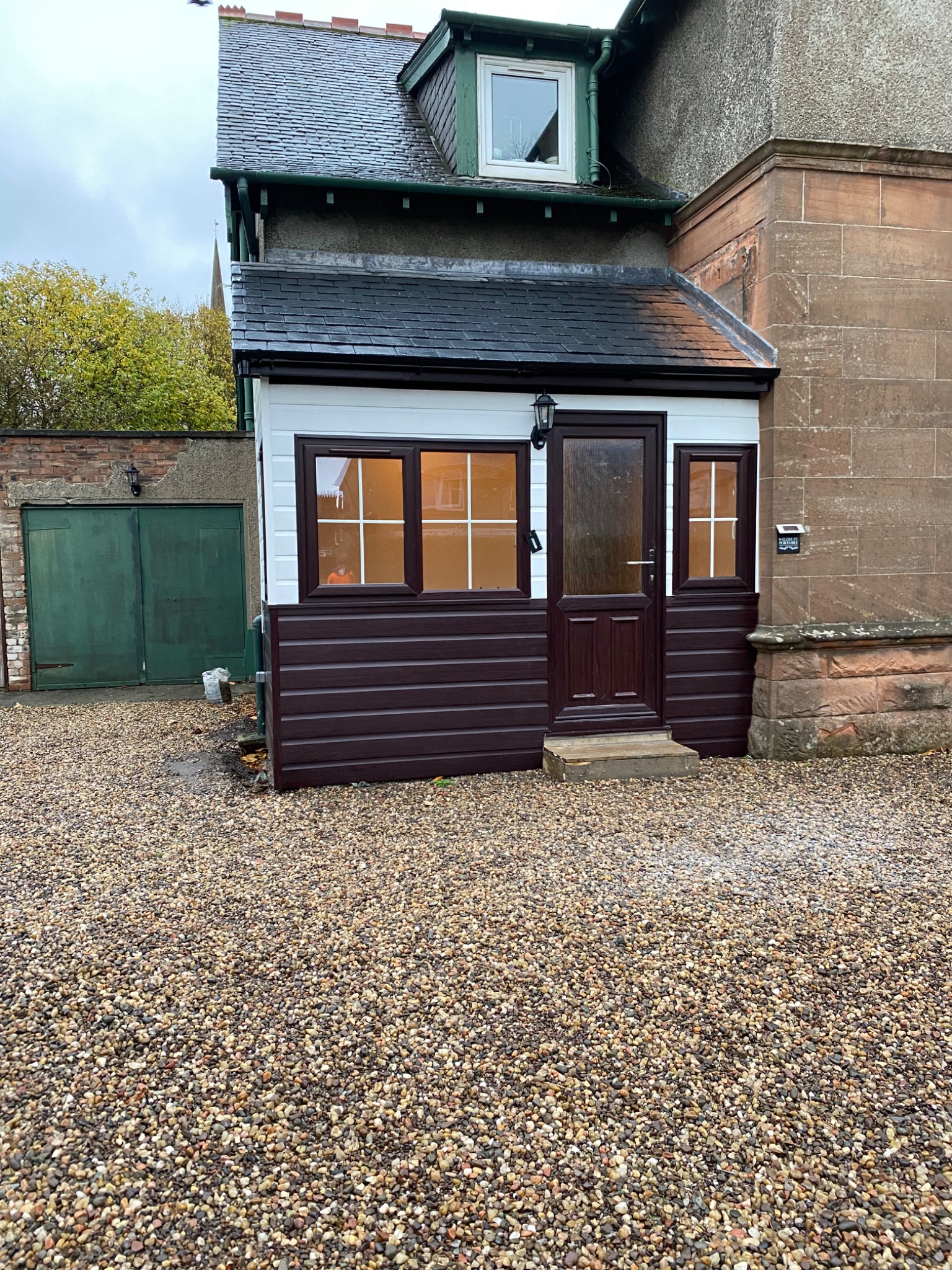 Zadaszone wejście do domu z brązowymi panelami i białymi ramami okiennymi, widok na garaż z zielonymi drzwiami i żwirowe podłoże.