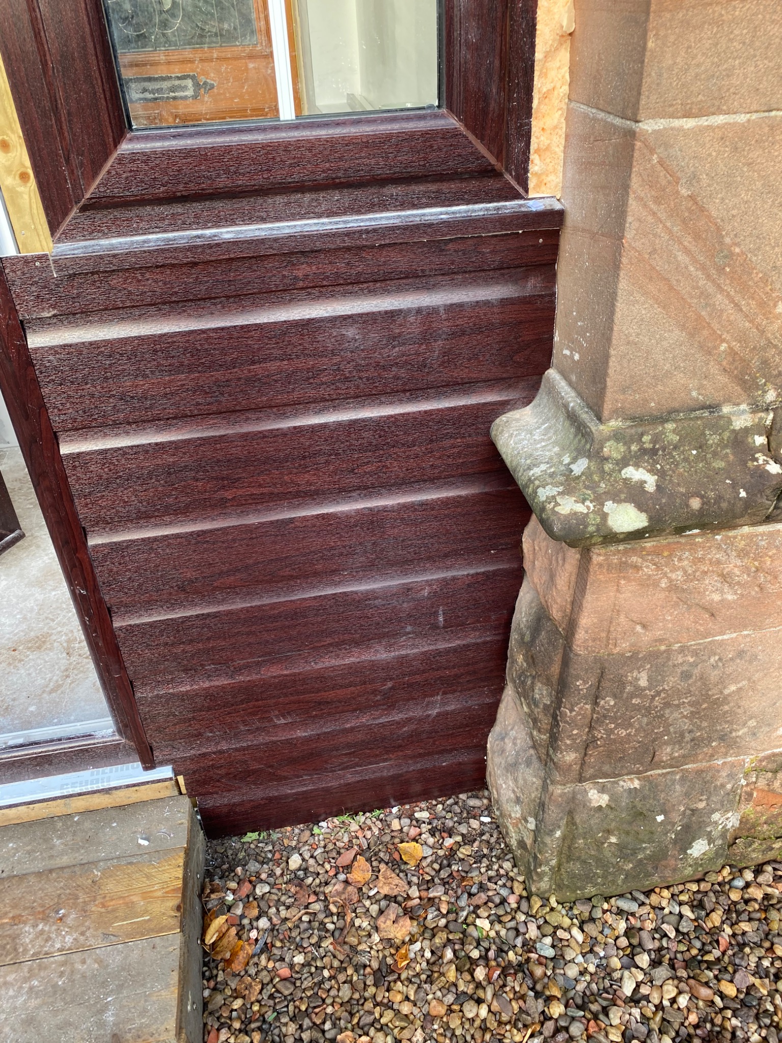 Fragment nowo zamontowanej, brązowej, drewnopodobnej elewacji bocznej, przylegającej do starego, kamiennego filaru budynku, z widocznym uszczelnieniem i drobnym żwirem u podstawy.